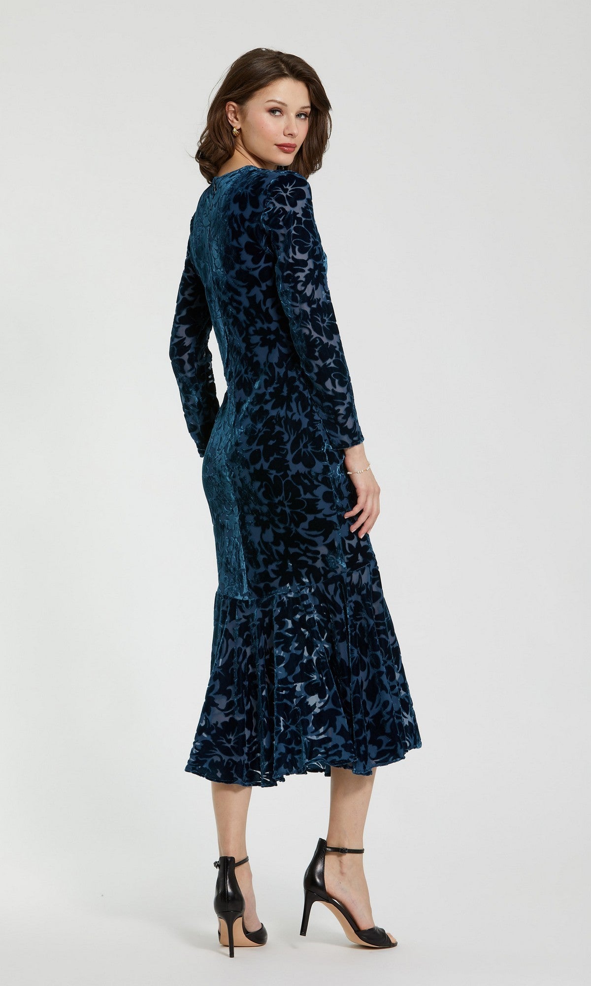 A woman stands against a plain light background, angled slightly away, wearing the Mac Duggal 81129—a fitted, long-sleeve blue burnout velvet midi party dress with a dark floral pattern and flared hem—styled with black ankle-strap heels for an elegant look.